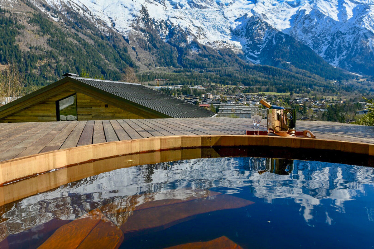 A luxurious outdoor hot spring pool with a wooden deck, reflecting snow-capped mountains and a small village in the background.