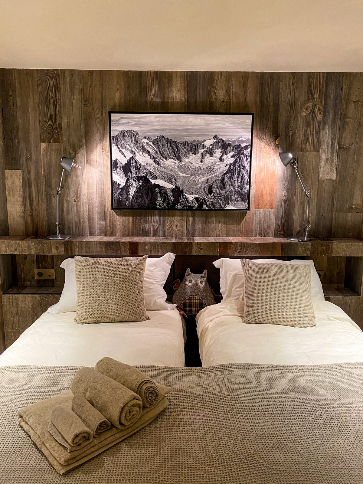 A styled mountain landscape wall art print in black and white, displayed above a double bed in a rustic bedroom with wood-paneled walls and bedside lamps.