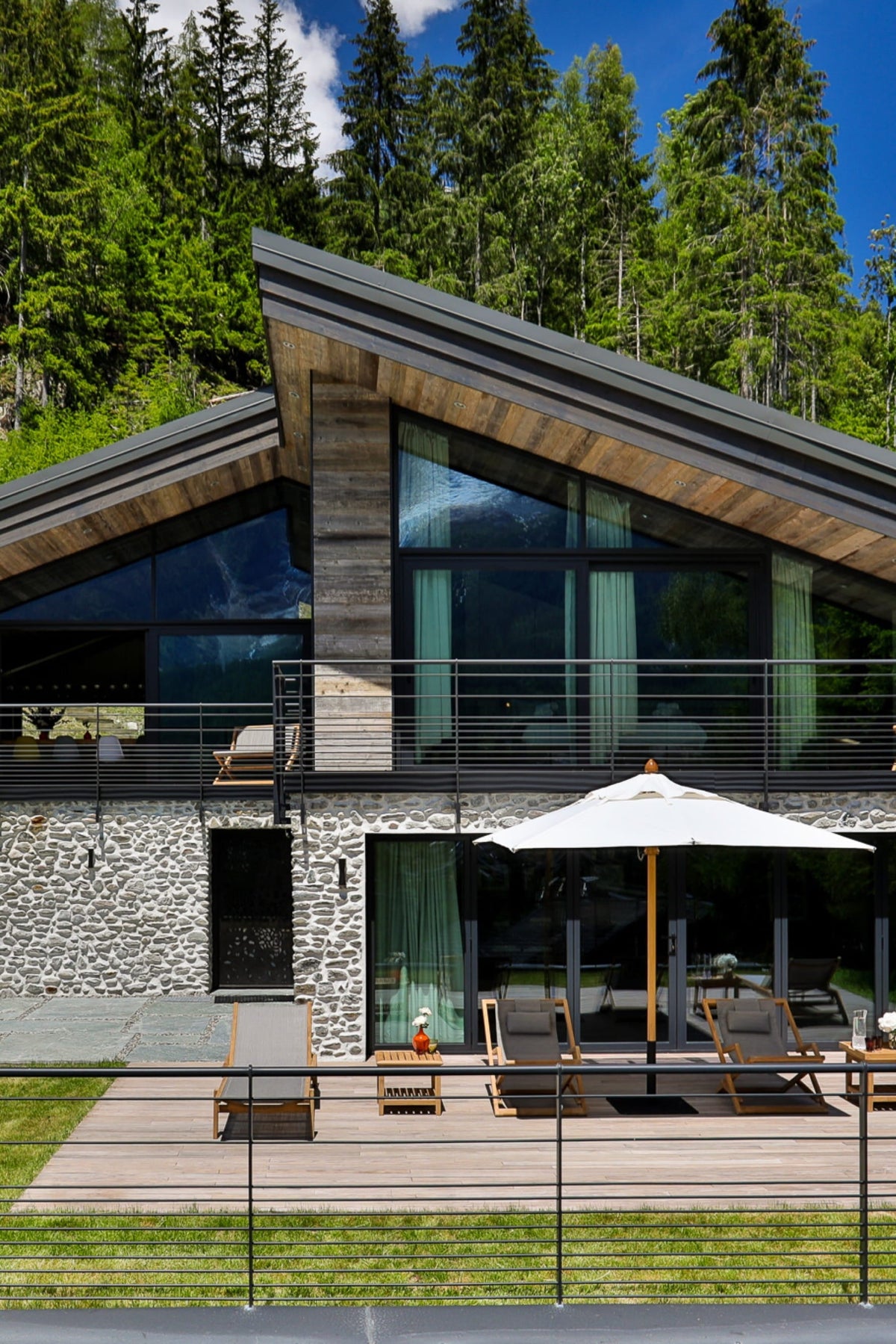 Modern mountain chalet with a stone and wood facade, large glass windows, a sloped roof, outdoor patio with lounge chairs and umbrella, surrounded by a forested hillside.