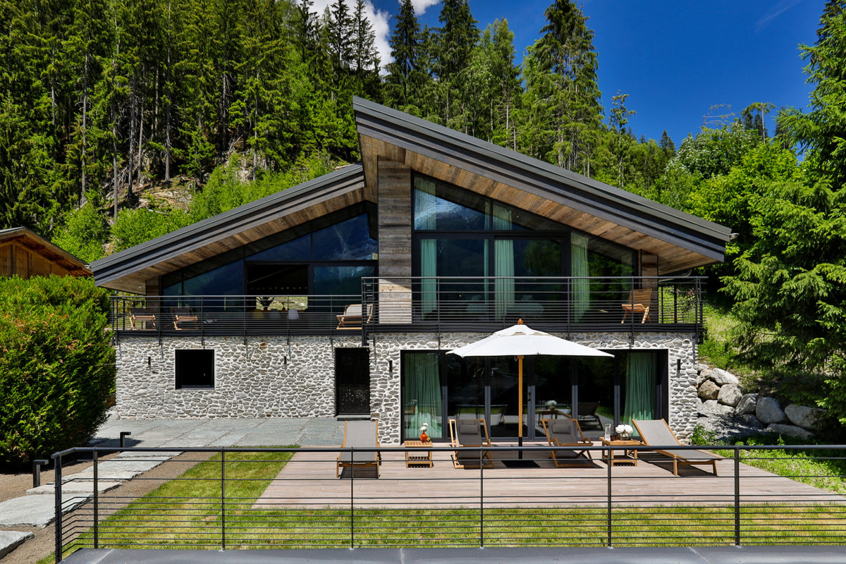 Modern mountain chalet with a stone and wood facade, large glass windows, a sloped roof, outdoor patio with lounge chairs and umbrella, surrounded by a forested hillside.