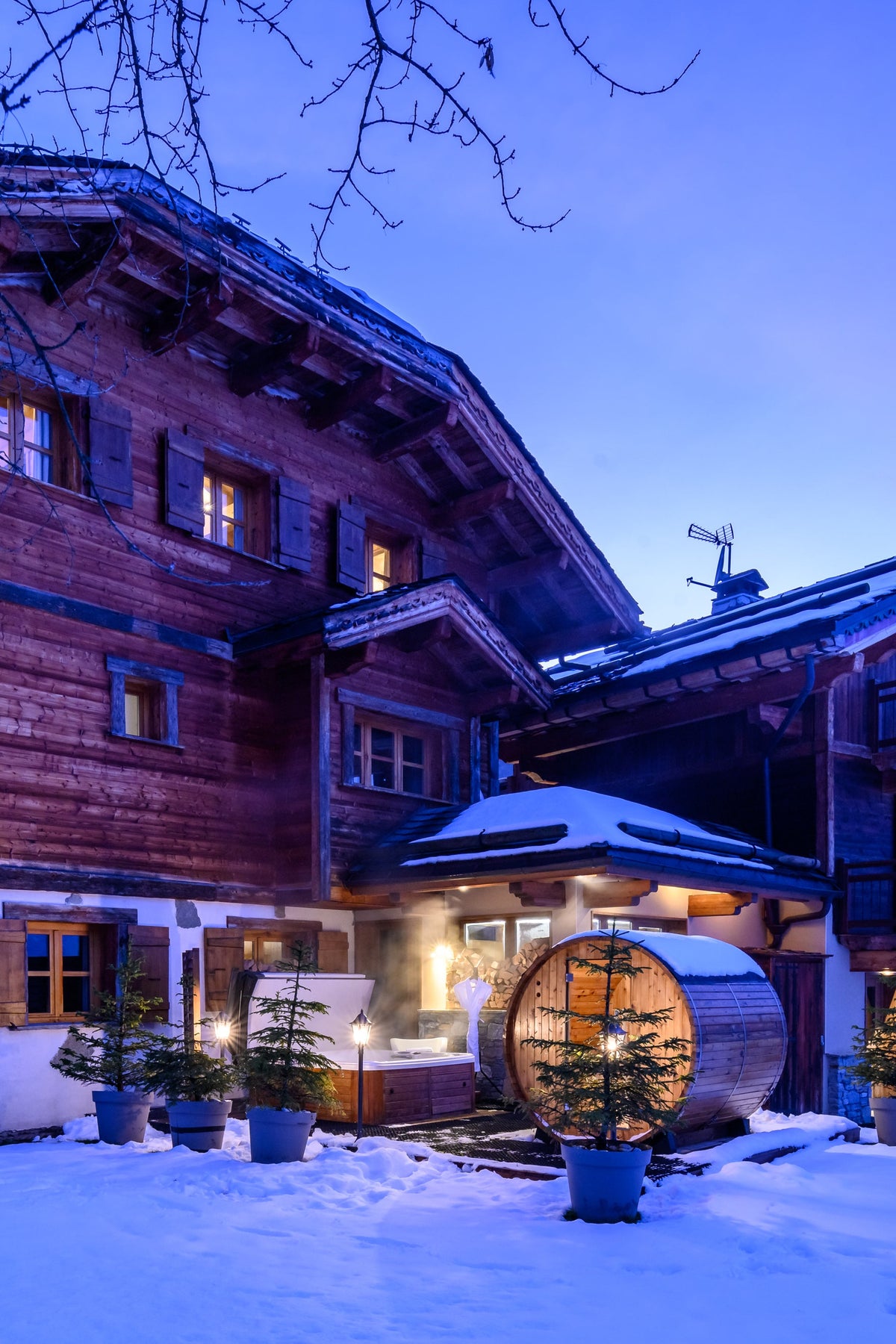 A large traditional alpine chalet with wooden walls and snow-covered roofs, lit warmly at dusk in a snowy mountain setting.