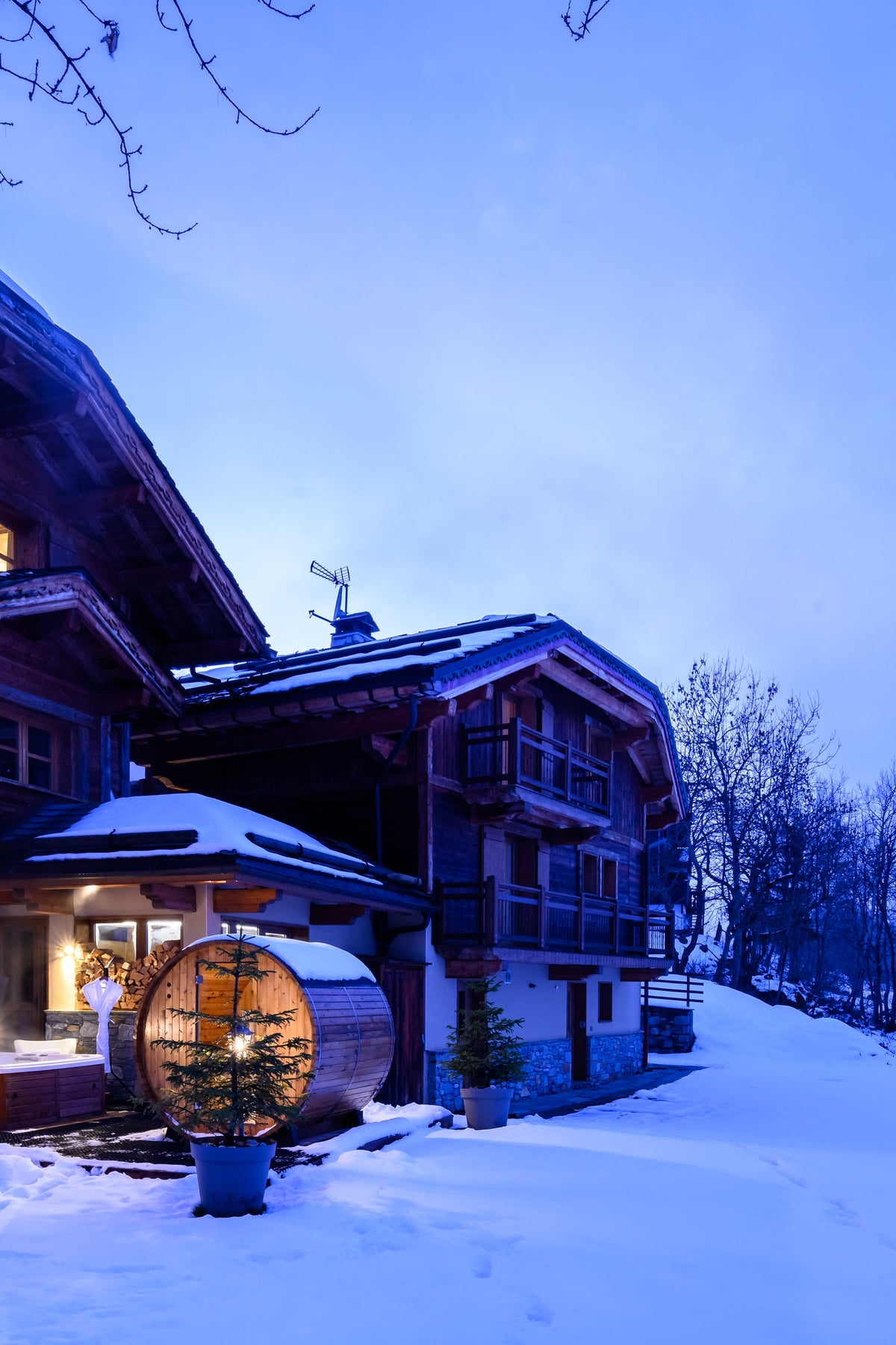 A large traditional wooden chalet in a snowy mountain setting at dusk, with warm lights glowing from the windows and a round wooden barrel structure in the snow in front.
