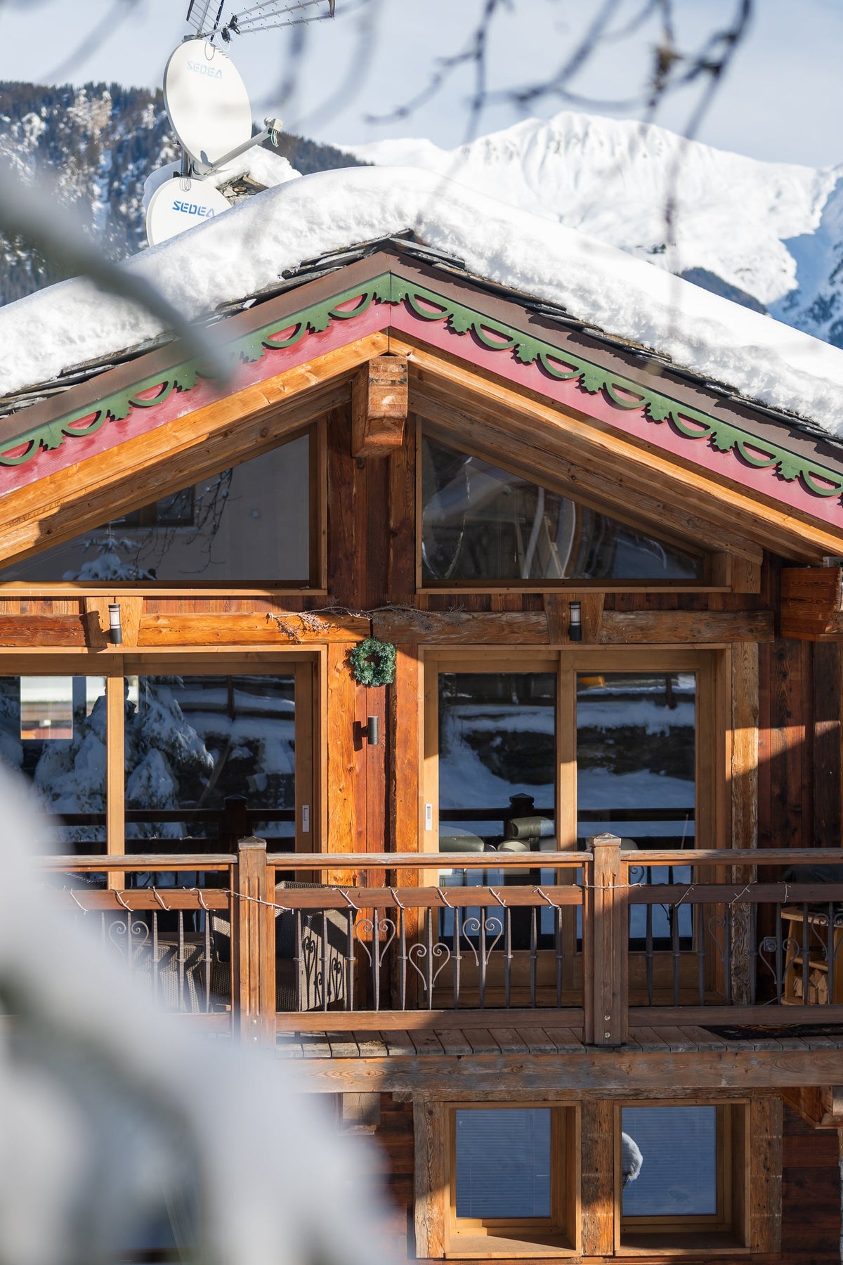 A rustic wooden chalet with snow-covered roof and balcony, set against a snowy mountain landscape with blurred tree branches in the foreground.