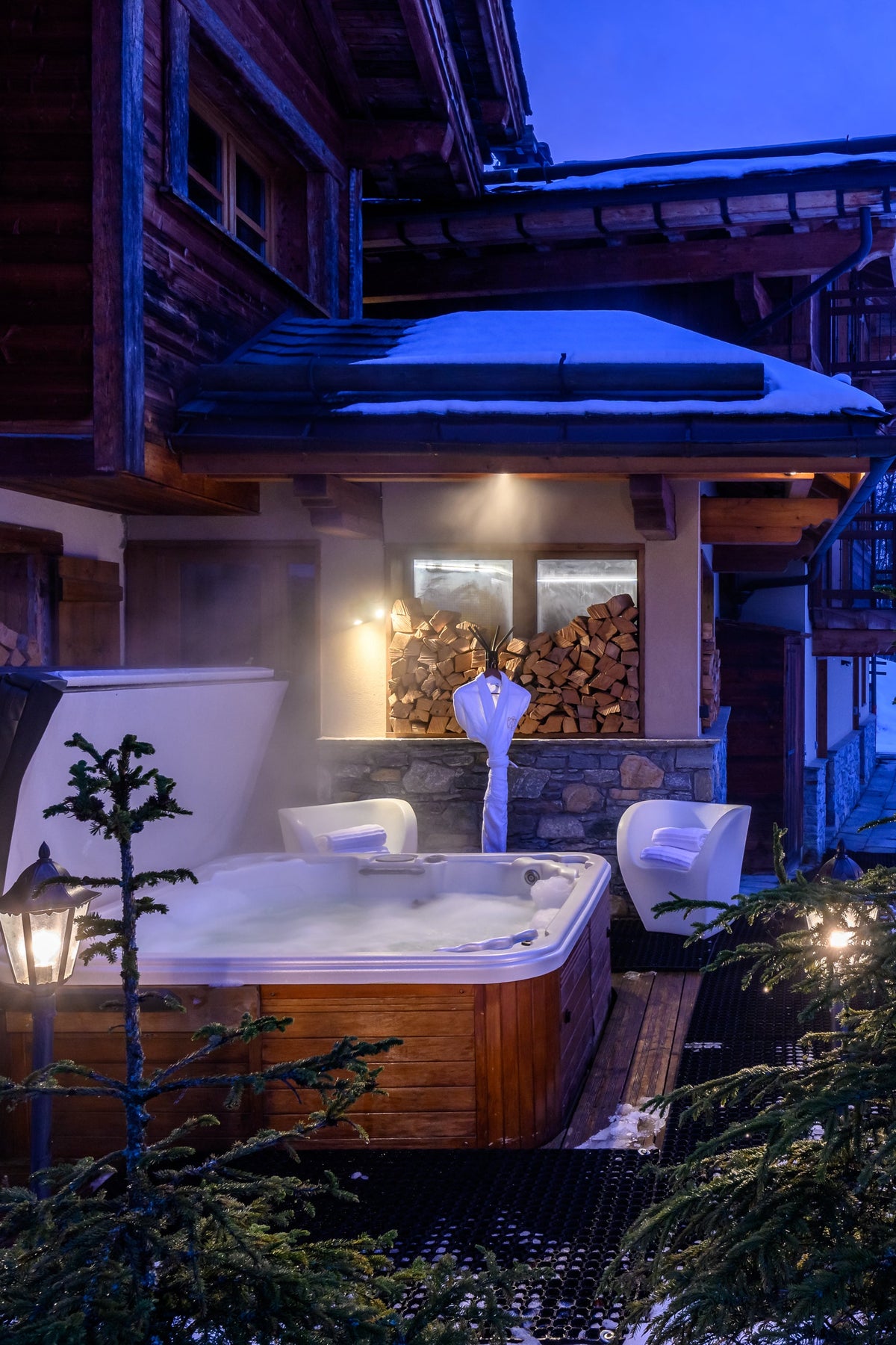 A wooden hot tub and a round wooden sauna are set outside a log cabin in a snowy winter landscape at dusk, with snow-covered ground, evergreen trees, and a blue evening sky.