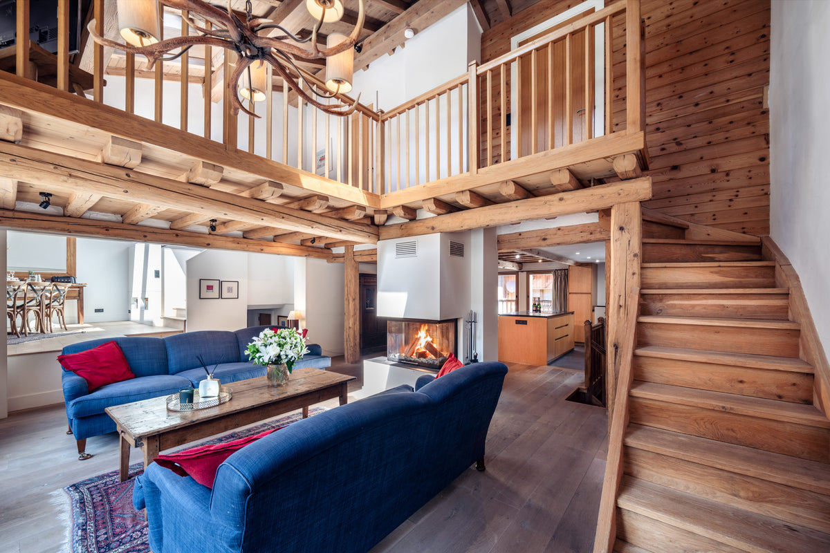 A rustic alpine chalet interior with a wooden staircase, loft balcony, blue sofas, a fireplace, and exposed wood beams.