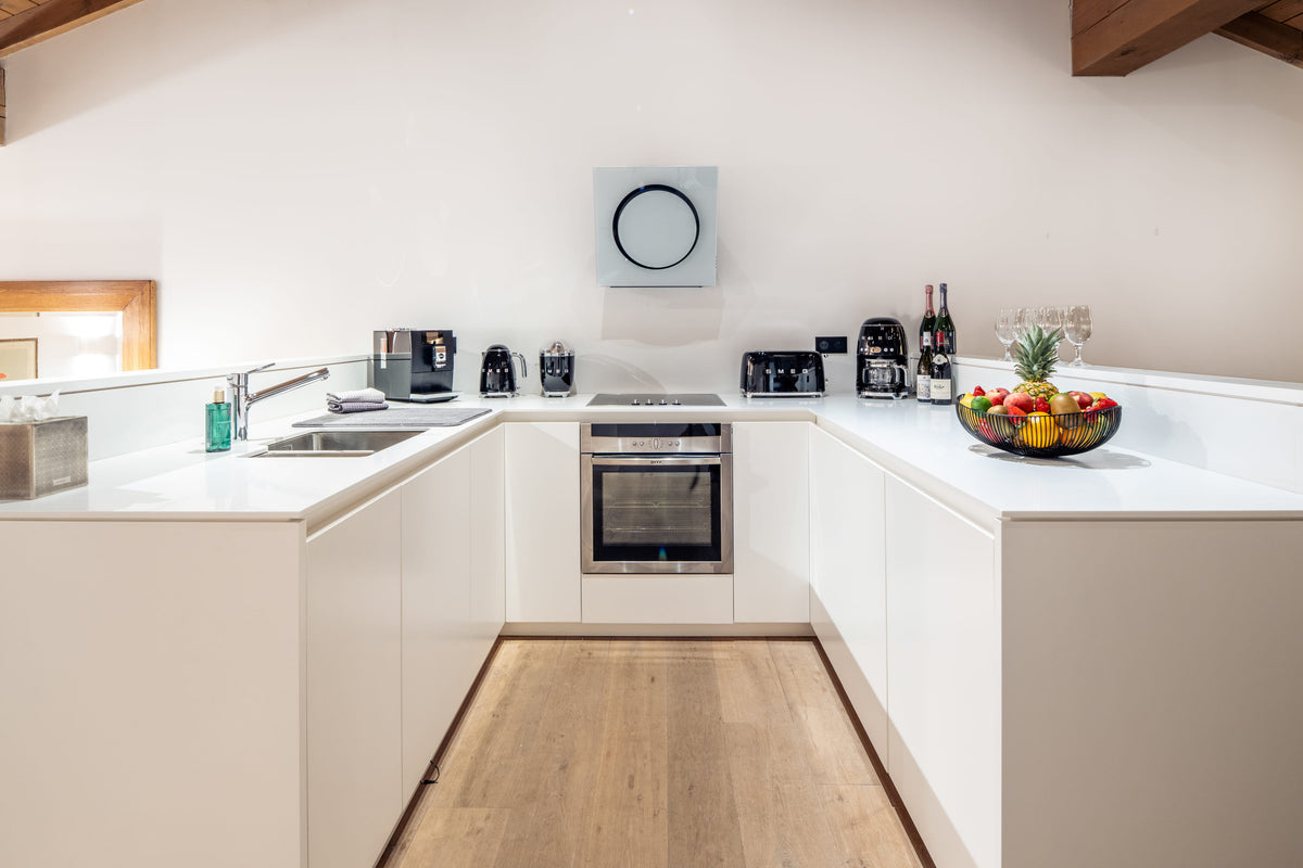 A modern U-shaped kitchen with white cabinets and countertops, a stainless steel oven, a sink, and a wooden floor. A bowl of fruit sits on the right counter.
