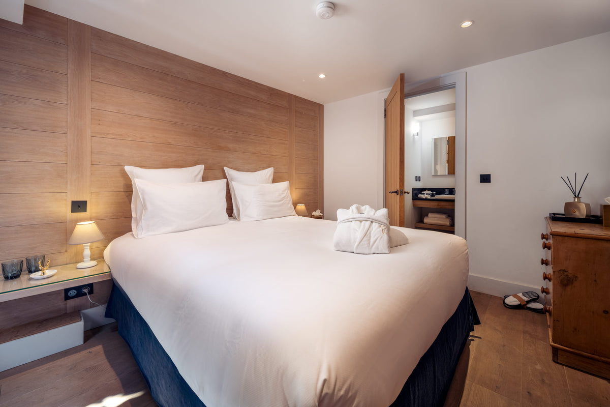 A modern bedroom featuring a large bed with white bedding and a dark blue skirt, set against a light wood-paneled headboard. The room includes a wooden nightstand with a lamp, a wooden dresser, and a visible bathroom area in the background.