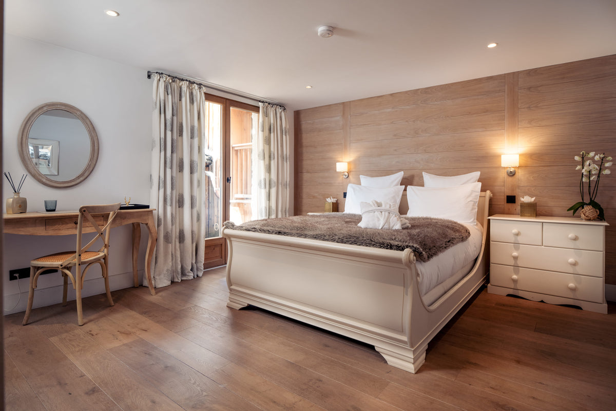 A beautifully styled bedroom featuring a cream-colored wooden sleigh bed with white linens and a brown faux-fur throw, set against a light wood-paneled wall. The room includes a matching cream nightstand, a wooden desk with a chair, a round mirror, and a large window with curtains.