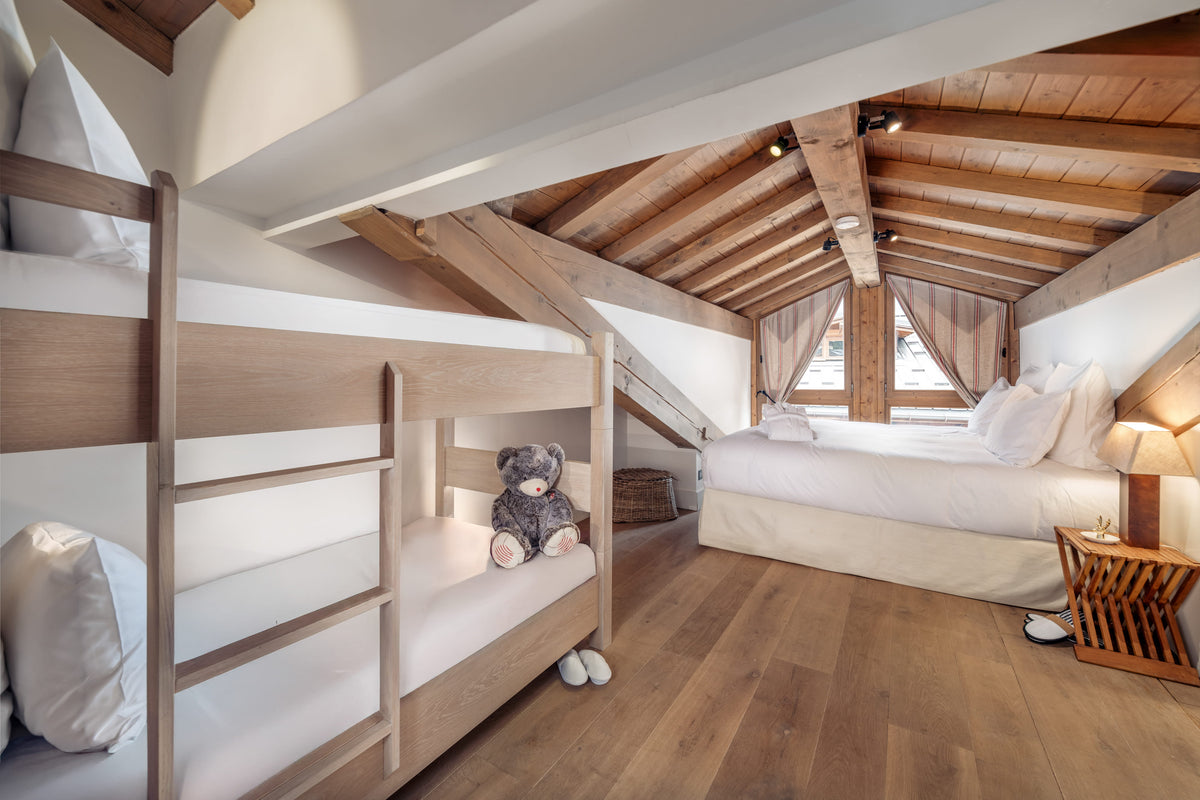 A cozy attic bedroom with a wooden bunk bed on the left and a single bed on the right, featuring a sloped wooden ceiling, warm wood floors, white bedding, and a small teddy bear on the bunk.