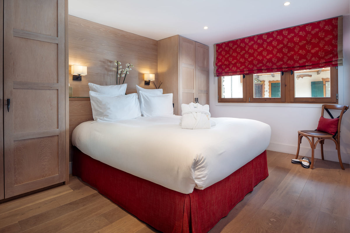 A double bed with white bedding and a red skirt in a modern bedroom with light wood furniture, a red patterned window valance, and a wooden chair.