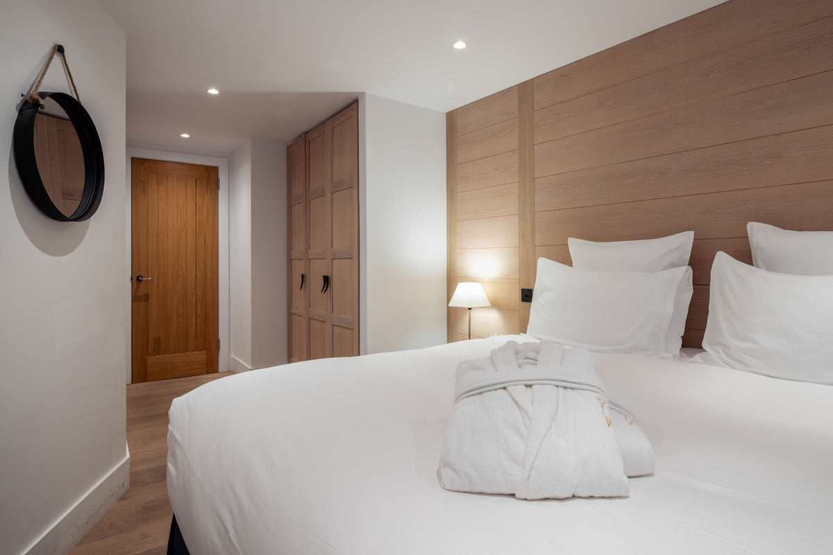 A modern bedroom featuring a neatly made bed with white linens and a folded white robe, set against a light wood headboard and matching wood wardrobes in a bright, contemporary room.
