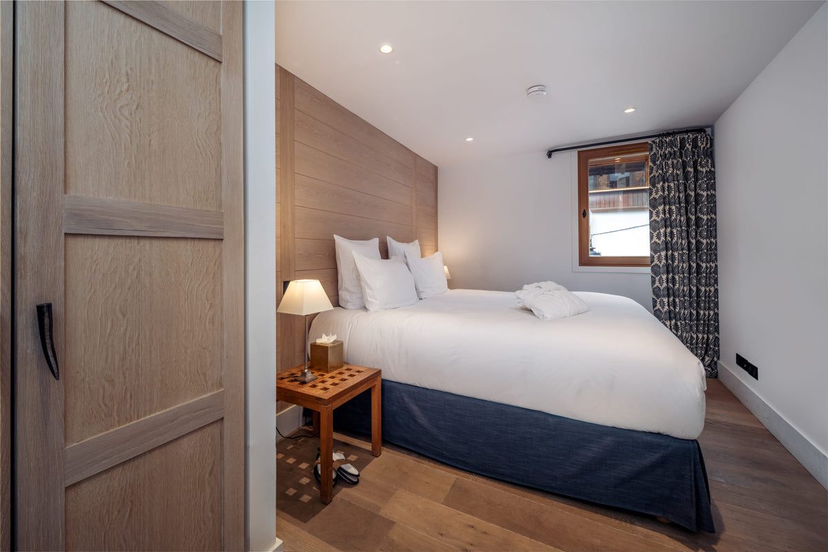 A modern bedroom featuring a neatly made bed with white bedding and a dark navy blue bed skirt, set against a light wood-paneled headboard. A matching wooden nightstand with a lit table lamp sits beside the bed.