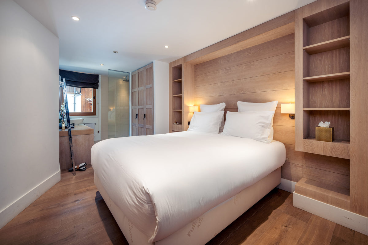 A modern bedroom featuring a large bed with white bedding, set against a light wood paneled headboard with built-in shelving and warm ambient lighting.
