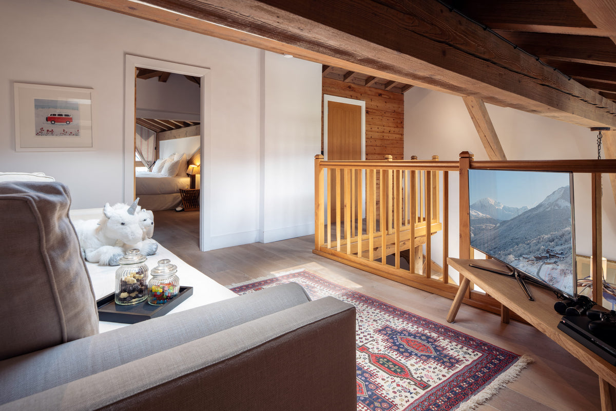 A cozy modern chalet living room featuring a gray fabric sofa, a patterned oriental rug, a wooden staircase railing, and a TV mounted on a wooden console. A white plush unicorn toy sits on the sofa next to an open doorway leading to a bedroom.