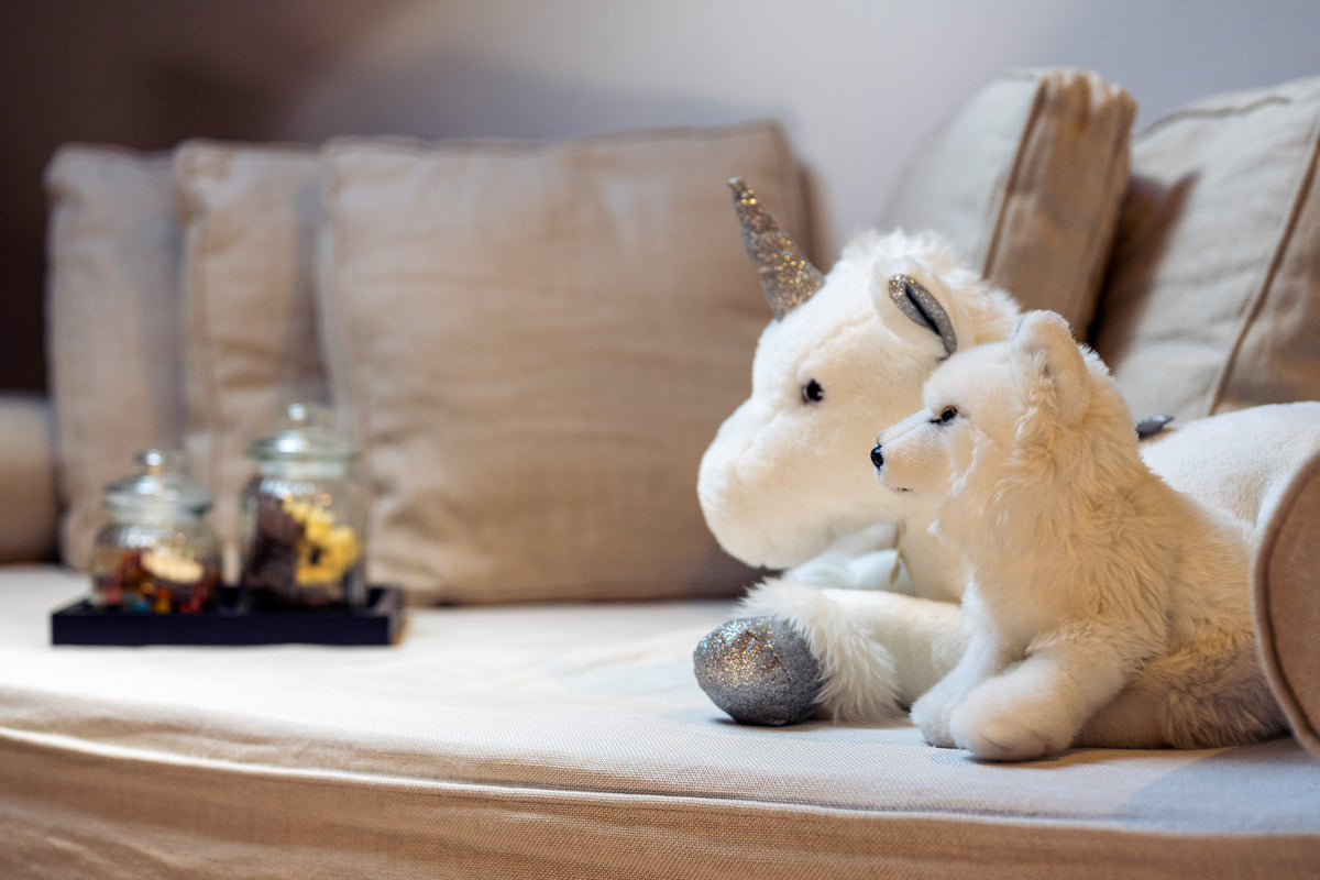 A plush white unicorn toy with a glittery silver horn and hooves resting on a beige sofa next to a smaller white plush toy, with soft pillows and decorative glass jars in the background.