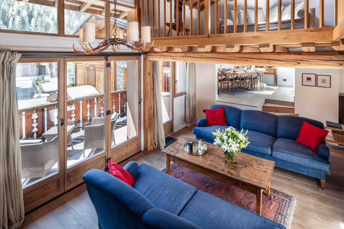 A bright, rustic chalet-style living room with two blue sofas, red accent pillows, a wooden coffee table, and large glass doors looking out onto a balcony and outdoor patio.