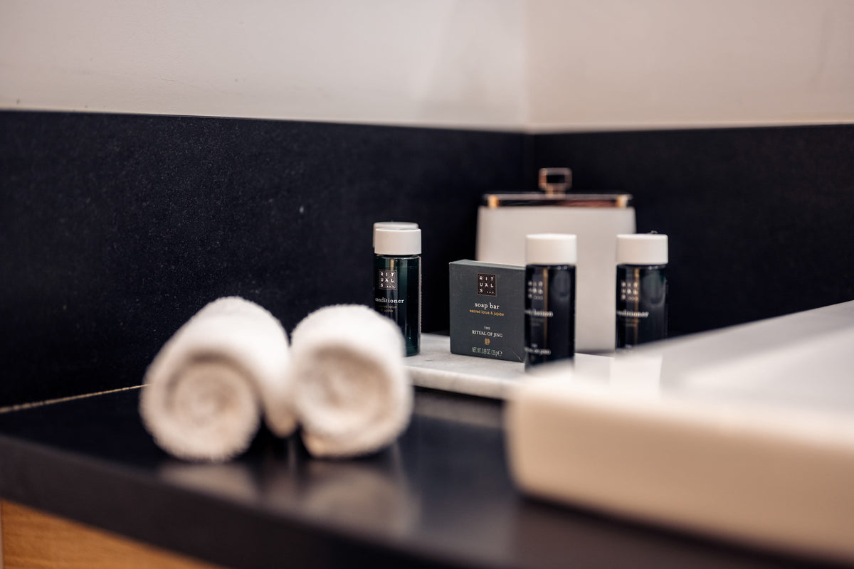 Hotel bathroom vanity with two rolled white towels and small toiletry bottles and a soap dish on a dark countertop.