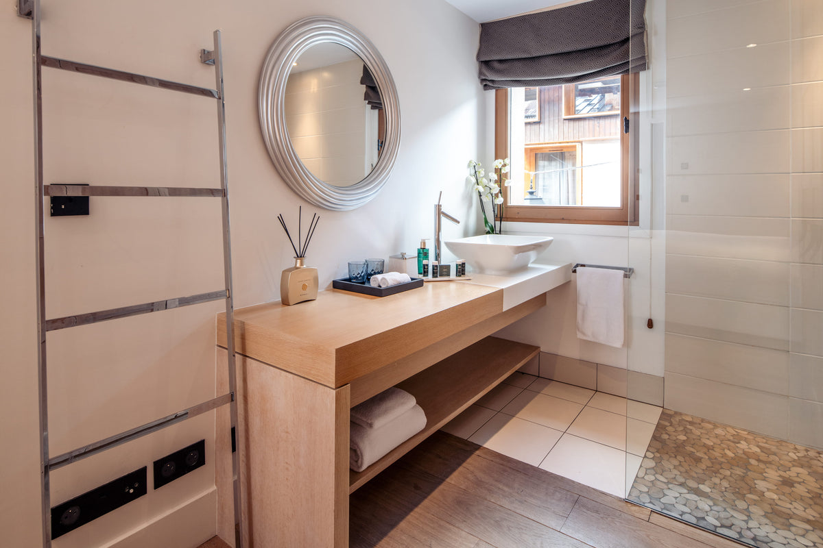 A modern bathroom vanity with a light wood console table, a white vessel sink, a round silver-framed mirror, and a walk-in shower area with white tiled walls.