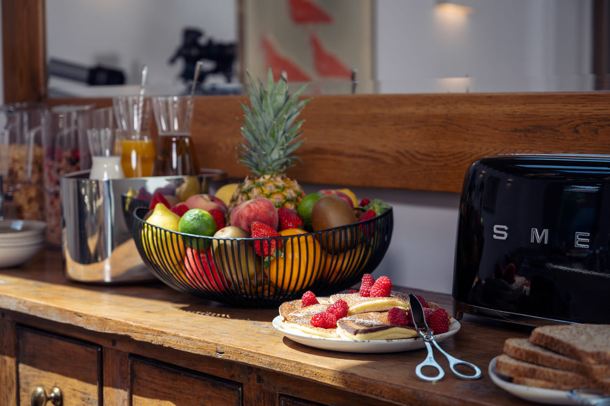 A breakfast buffet setup on a wooden counter featuring a black wire fruit basket with a pineapple, fresh strawberries, oranges, and other fruits, alongside a plate of pancakes topped with raspberries, a plate of toast, a metallic ice bucket, and a black SMEG toaster.