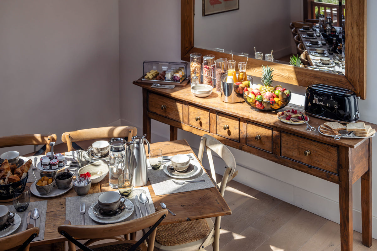A styled breakfast scene in a dining room featuring a wooden console table with a mirror, a dining table set with plates, cups, and bread, and a black toaster on the console.
