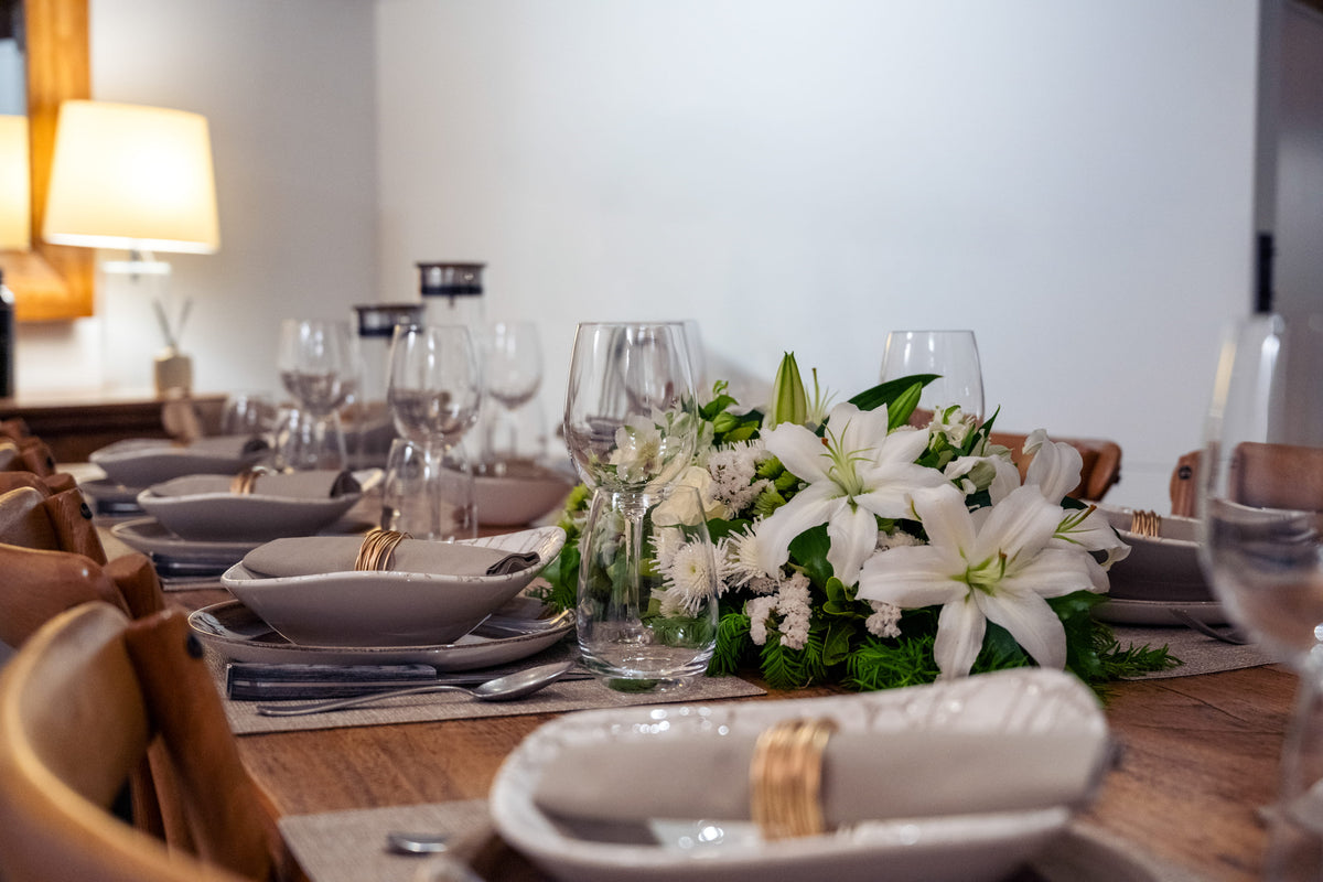 A beautifully set dining table with white lilies and green foliage centerpiece, white ceramic plates, gold ring napkins, and clear wine glasses in a warmly lit dining room.