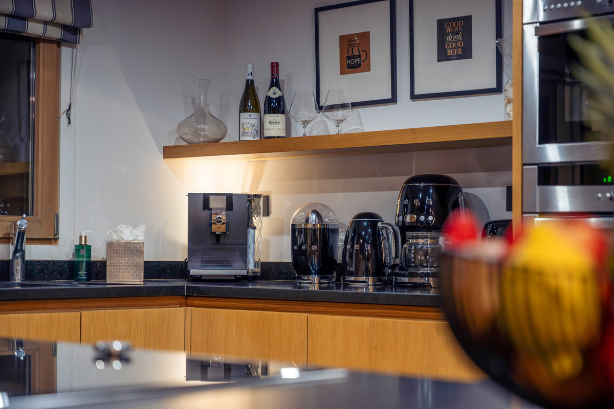 A modern kitchen countertop setup with black appliances including a coffee machine, kettle, and blender, set against light wood cabinetry and a white wall with framed art.