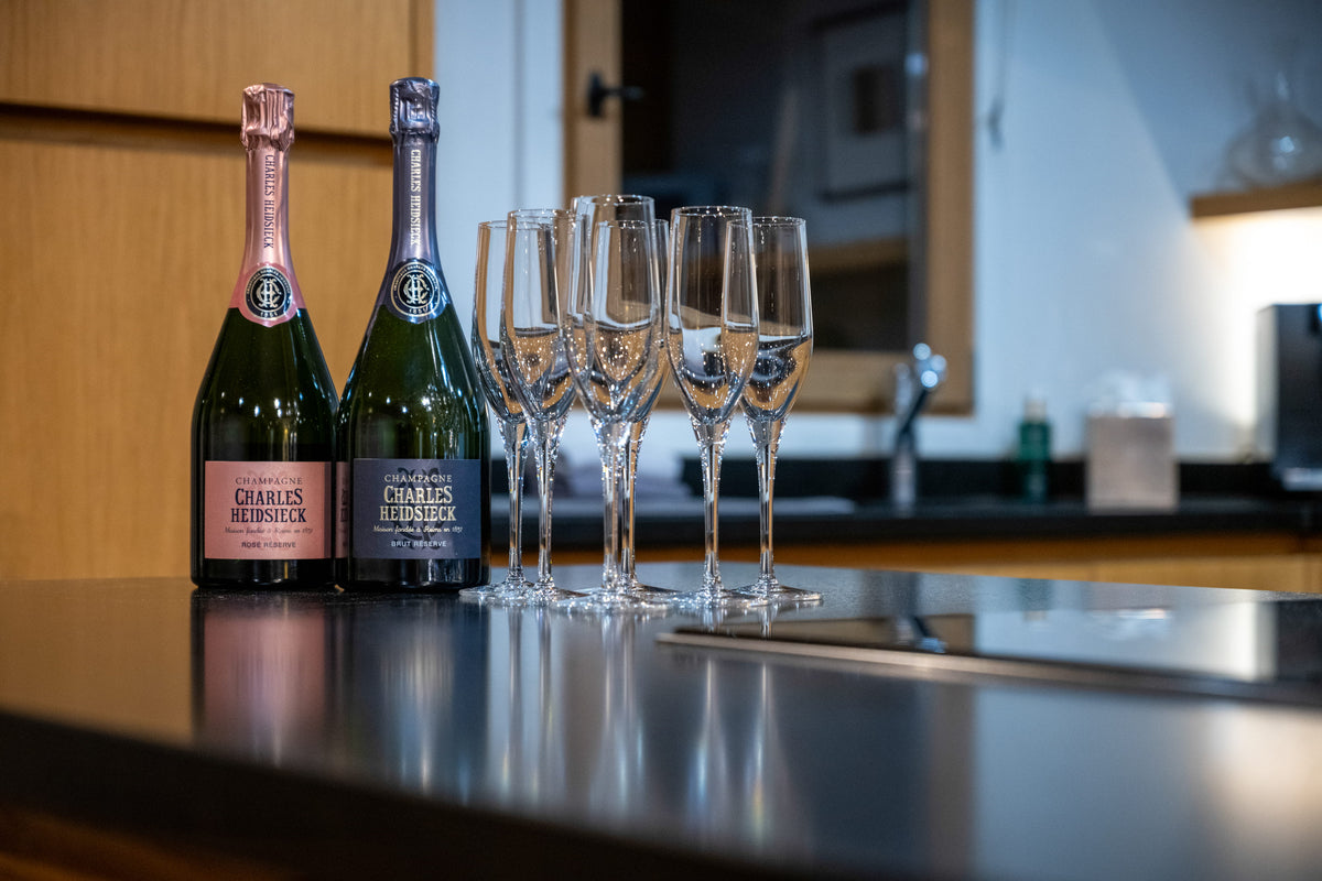 Two bottles of Charles Heidsig Brut Champagne and four clear champagne flutes arranged on a dark countertop in a kitchen setting.