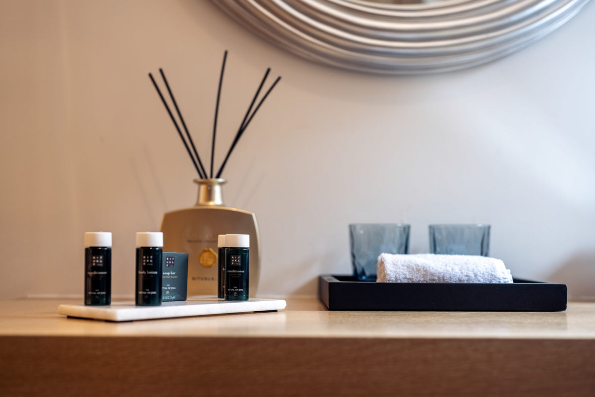 Four small dark travel-sized bottles with white caps arranged on a white tray on a wooden shelf, alongside a reed diffuser and a black tray holding two blue-gray glasses and a white towel.
