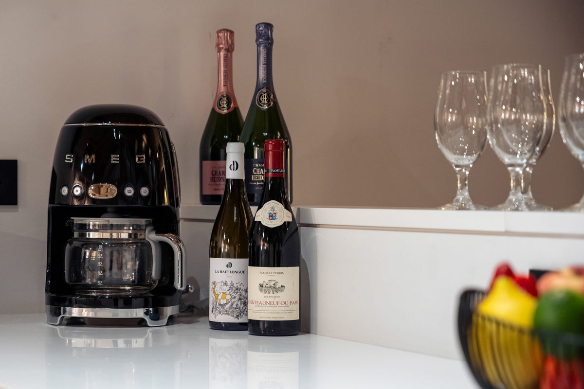 A black SMEG coffee machine sits on a white countertop next to several wine bottles and clear wine glasses on a white shelf, with a fruit bowl blurred in the foreground.