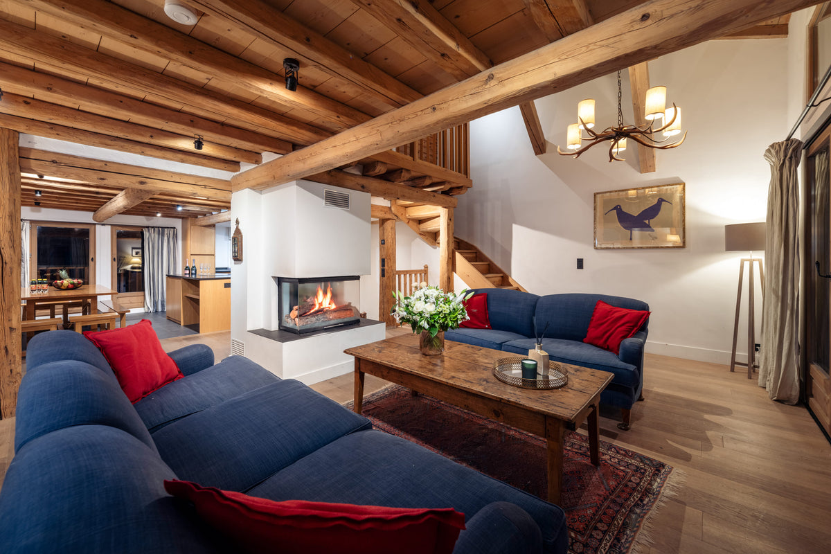 A cozy, rustic chalet-style living room featuring a large blue sectional sofa with red accent pillows, a matching blue sofa, a wooden coffee table, a fireplace, and exposed wooden ceiling beams.