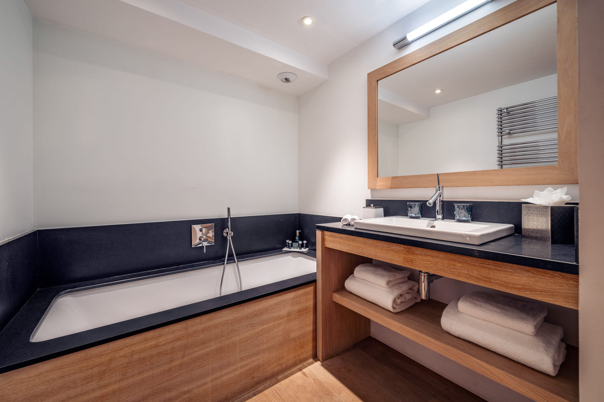 Modern bathroom interior featuring a built-in bathtub with a dark navy surround and wood base, a matching wood vanity with a white sink, large mirror, and folded white towels on open shelves.