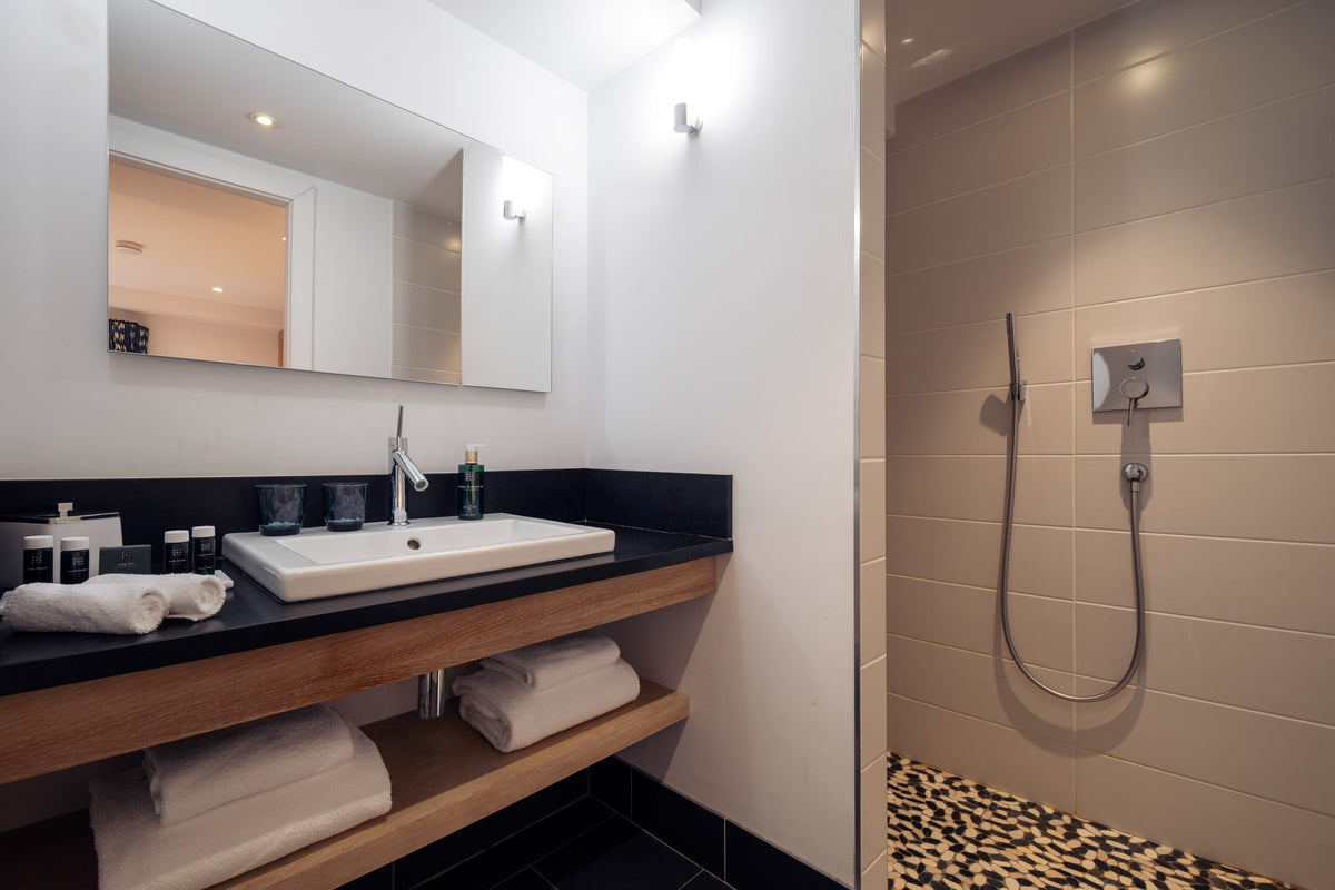 Modern bathroom vanity with a white rectangular sink, chrome faucet, black countertop, open wood shelving with folded white towels, a large wall mirror, and an adjacent tiled shower area with a leopard-print floor.