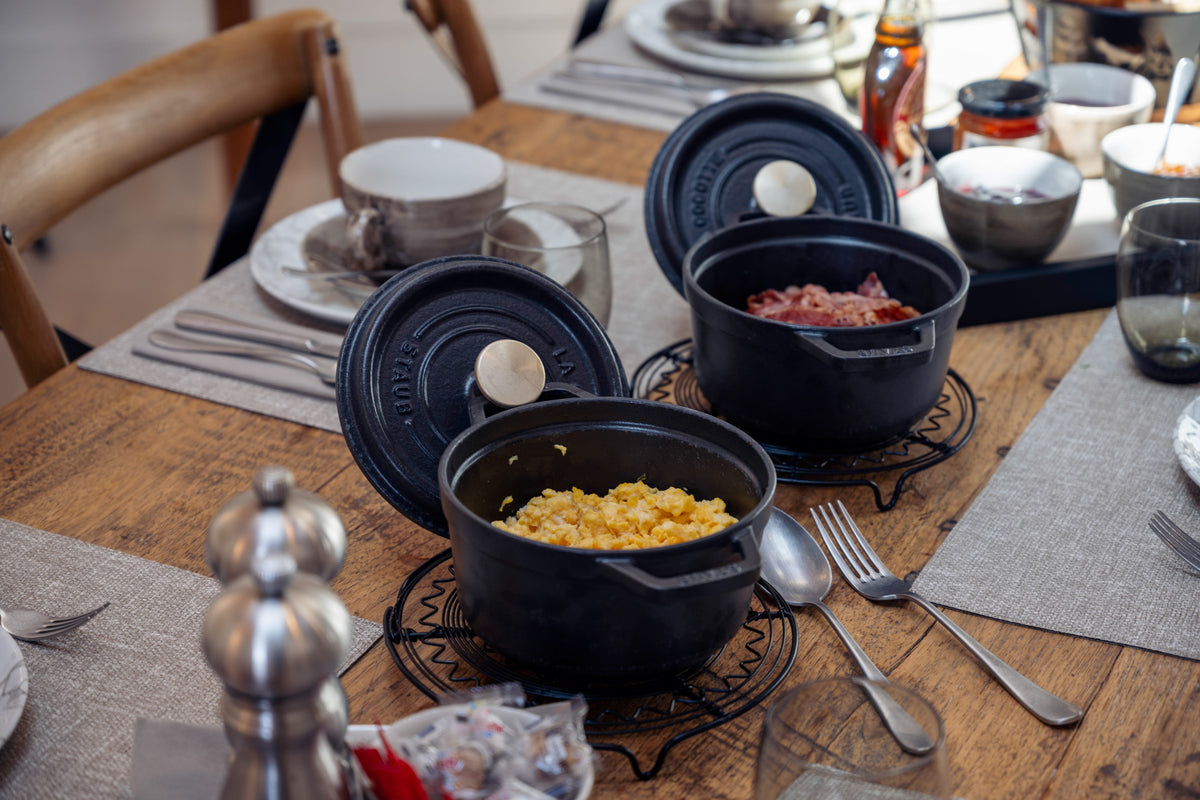 Two black cast-iron Dutch ovens on a wooden dining table, one filled with yellow rice and the other with pink food, surrounded by dining tableware.