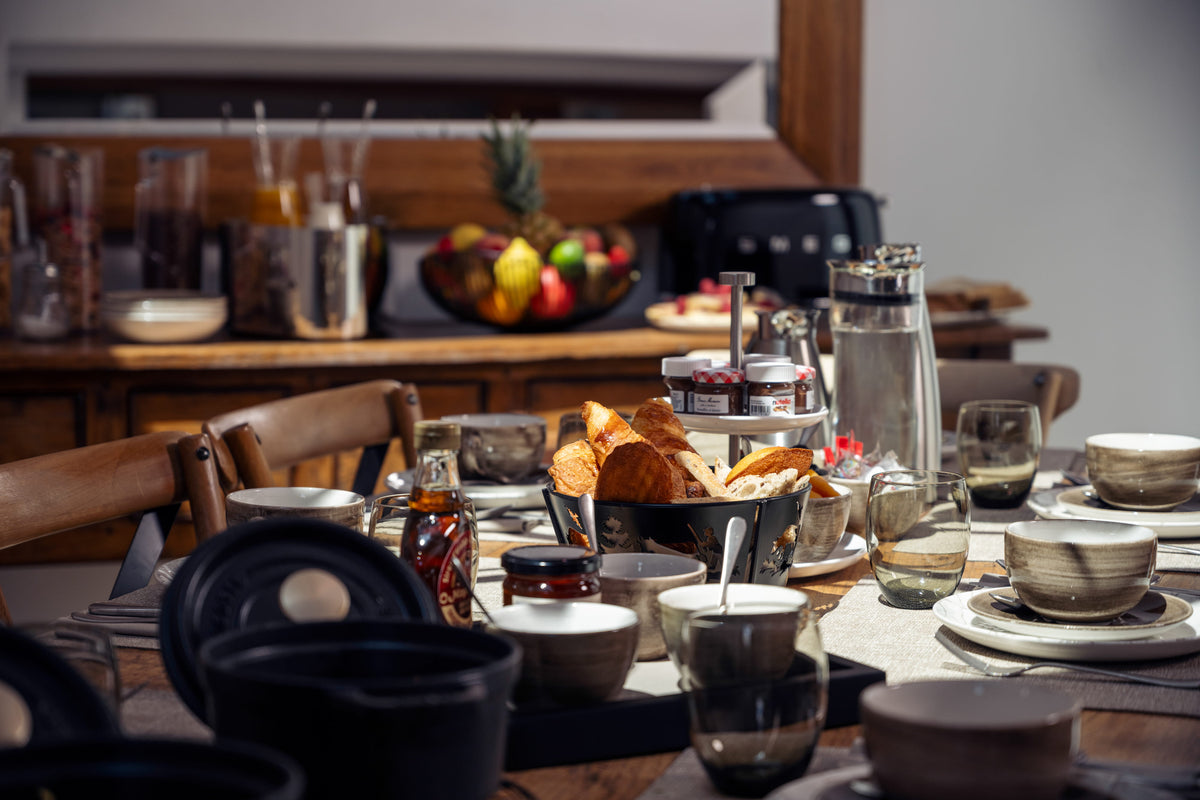 A styled dining table set with ceramic bowls, plates, glassware, and a basket of bread in a warm, rustic kitchen setting.