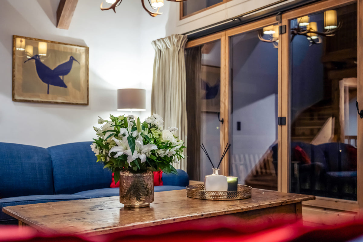 A cozy living room interior with a blue sofa, a rustic wooden coffee table holding a vase of white flowers and a reed diffuser, sliding glass doors, and framed wall art.