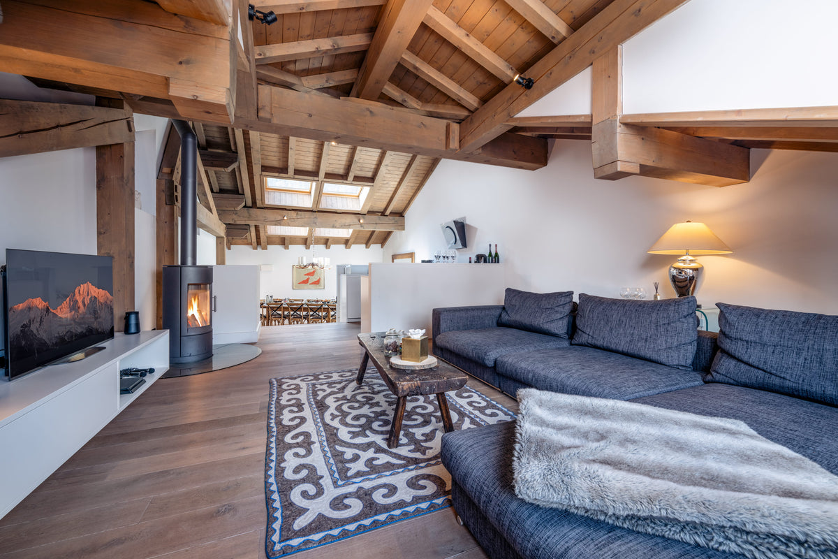 A cozy modern living room with a dark blue sectional sofa, a black wood-burning stove, a patterned rug, and a high sloped ceiling with exposed wooden beams.