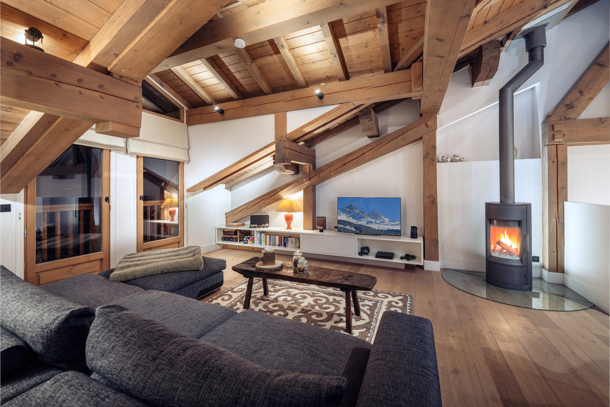 A cozy modern chalet living room featuring a dark gray sectional sofa, a rustic wooden coffee table, a patterned rug, a black wood-burning stove with a glass wind guard, and a low media console with a television. The room has a high sloped ceiling with prominent exposed wooden beams and light wood flooring.