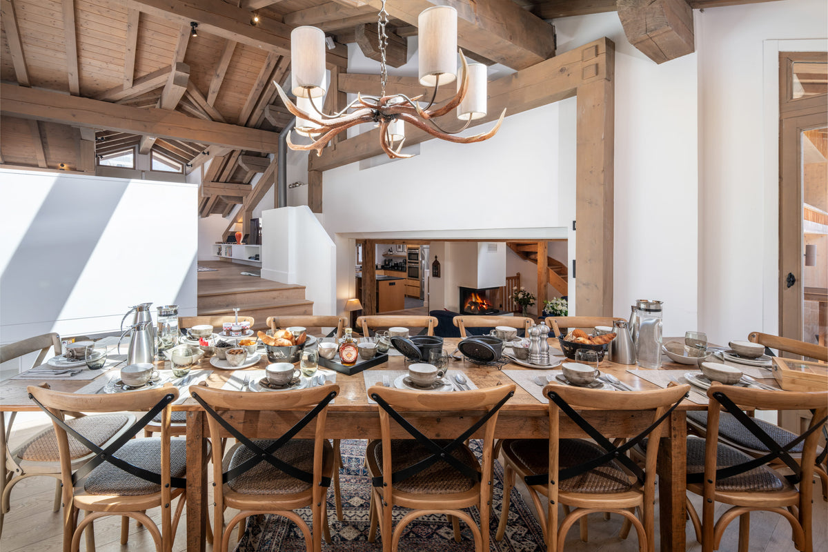 A rustic wooden dining table set with plates, cutlery, and glassware, surrounded by matching wooden chairs in a bright, modern chalet-style dining room with exposed wood beams and a chandelier.