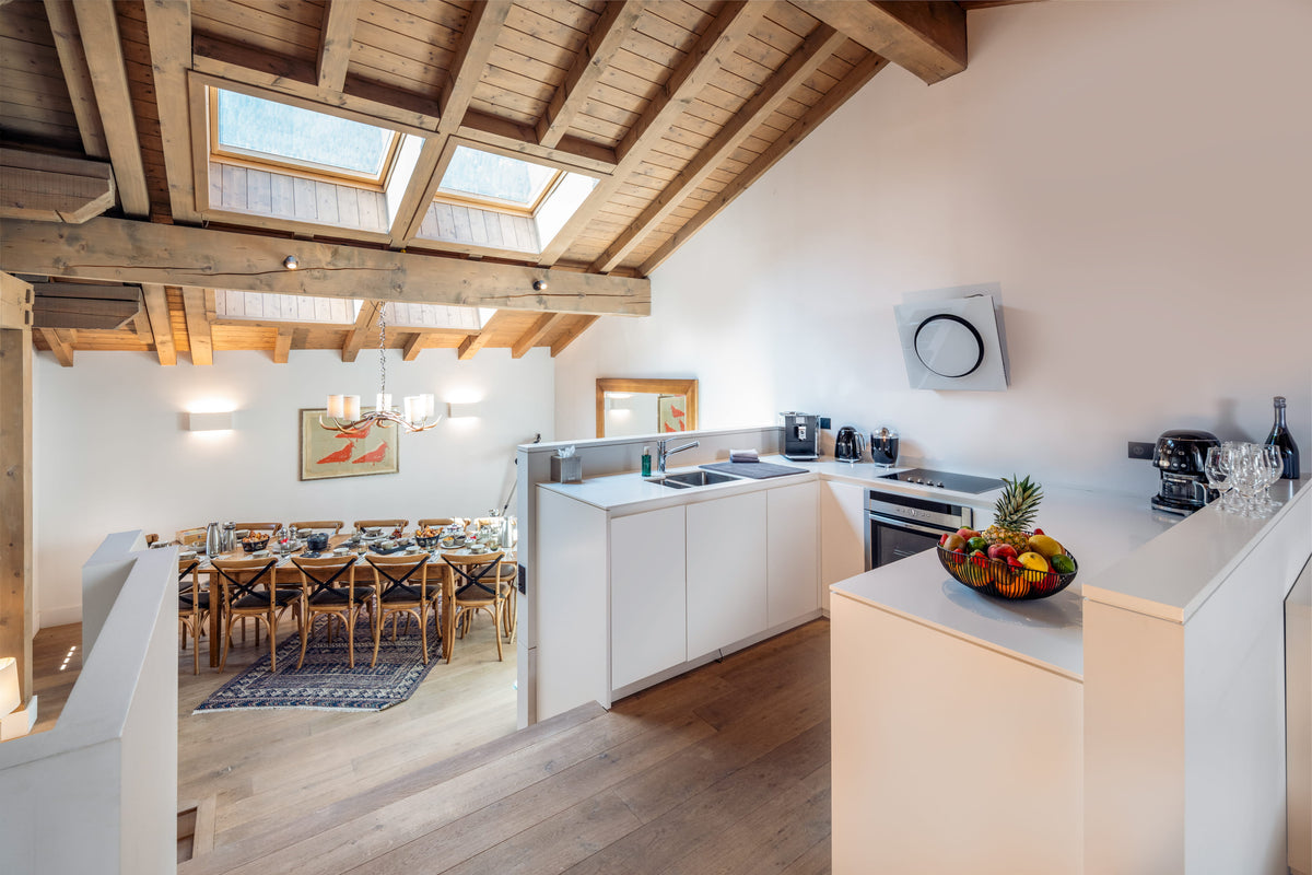 A bright, modern kitchen and dining area in a loft-style apartment with exposed wooden ceiling beams and skylights. The kitchen features white cabinets, a countertop with a fruit bowl, and a built-in oven.