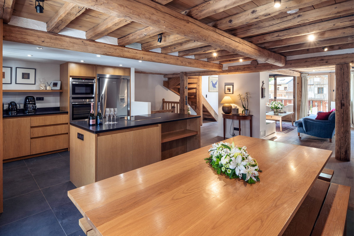A modern rustic kitchen island with a black countertop and wood cabinetry, set within a large open-plan chalet featuring exposed wooden ceiling beams, a wooden dining table with flowers, and a living area in the background.
