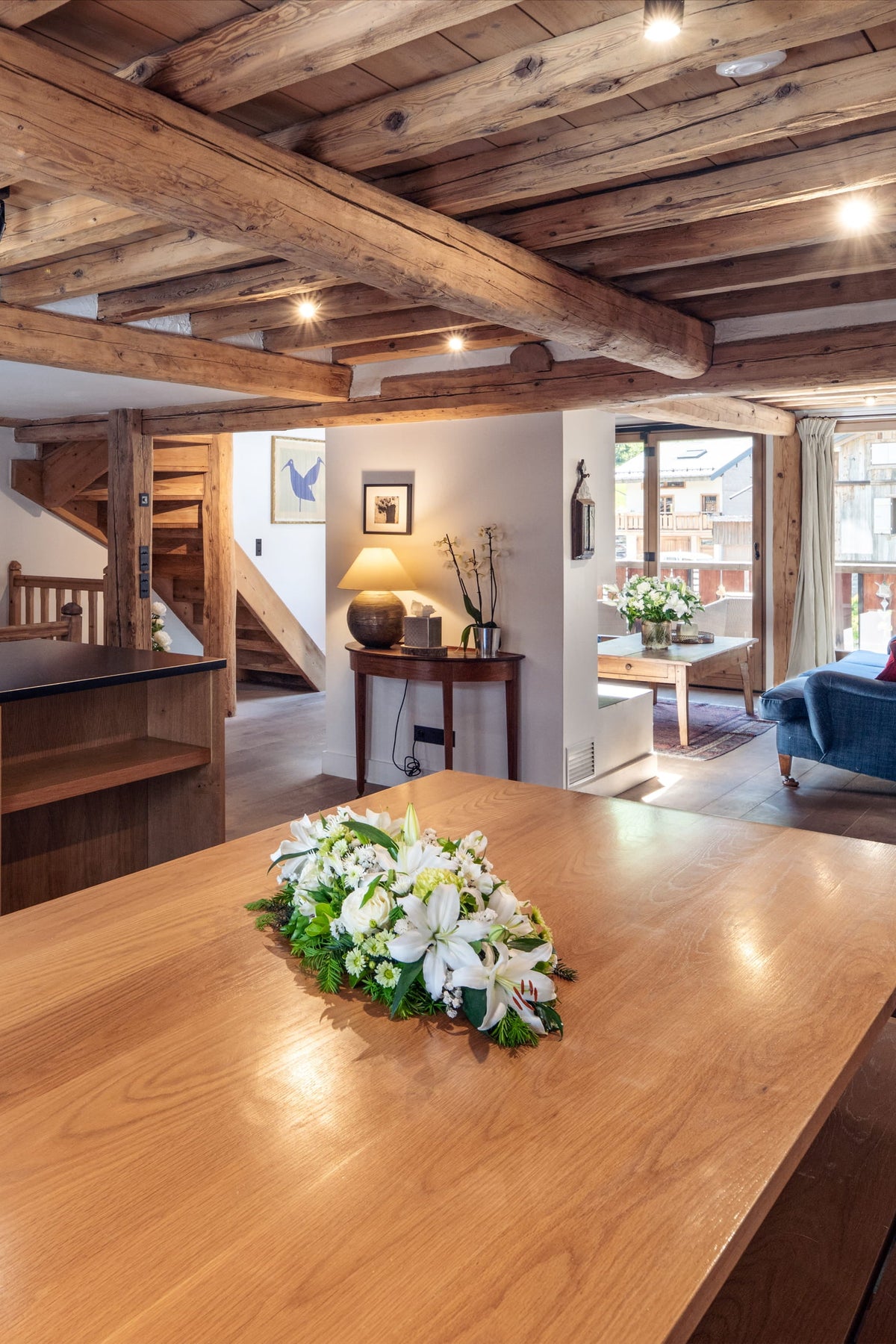 A modern rustic kitchen island with a black countertop and wood cabinetry, set within a large open-plan chalet featuring exposed wooden ceiling beams, a wooden dining table with flowers, and a living area in the background.
