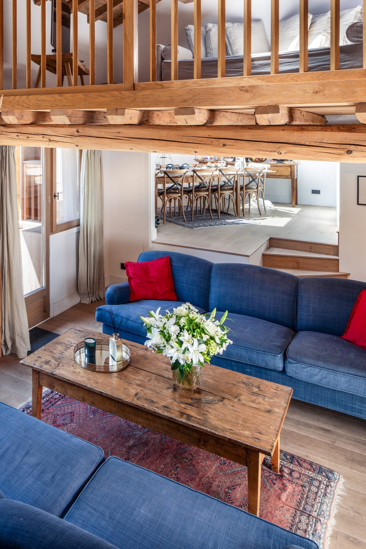 A bright, rustic chalet-style living room with two blue sofas, red accent pillows, a wooden coffee table, and large glass doors looking out onto a balcony and outdoor patio.