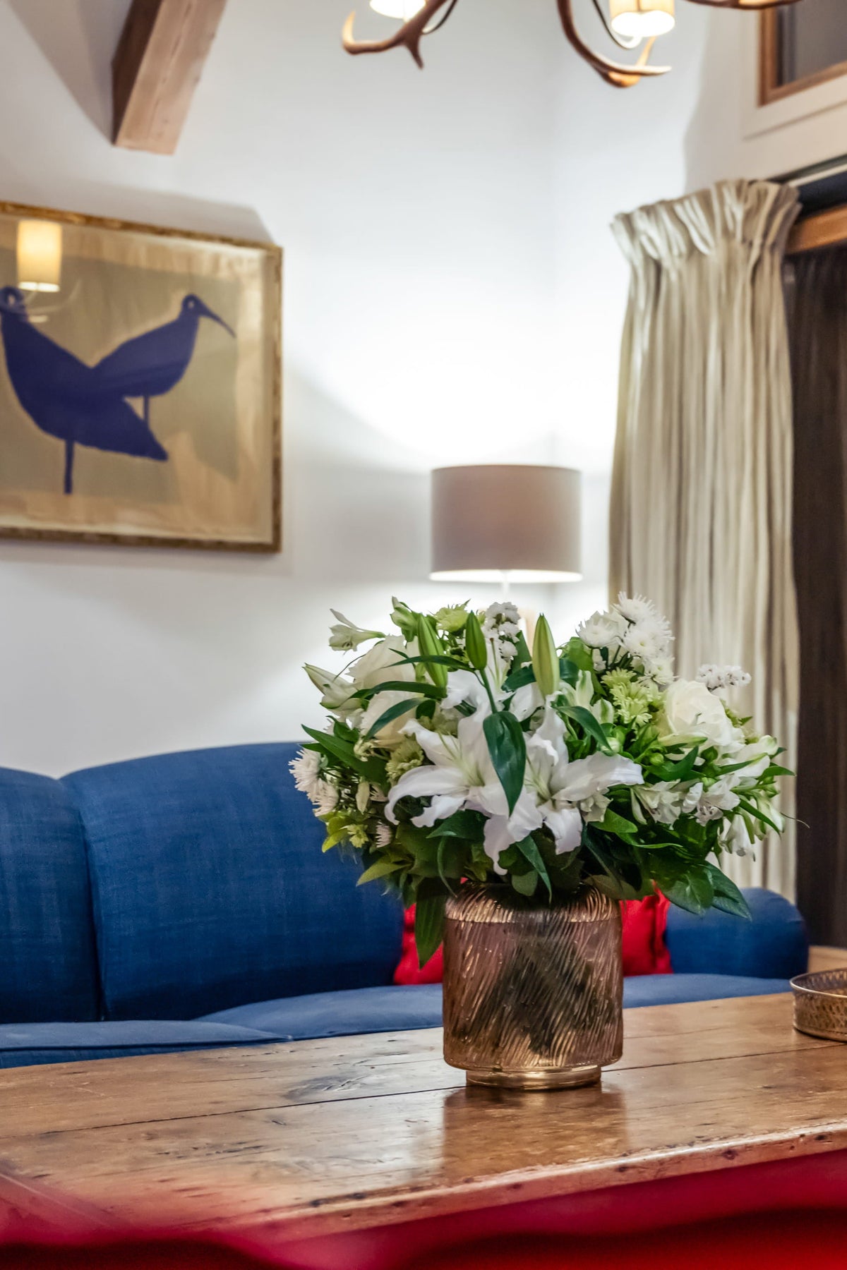 A cozy living room interior with a blue sofa, a rustic wooden coffee table holding a vase of white flowers and a reed diffuser, sliding glass doors, and framed wall art.