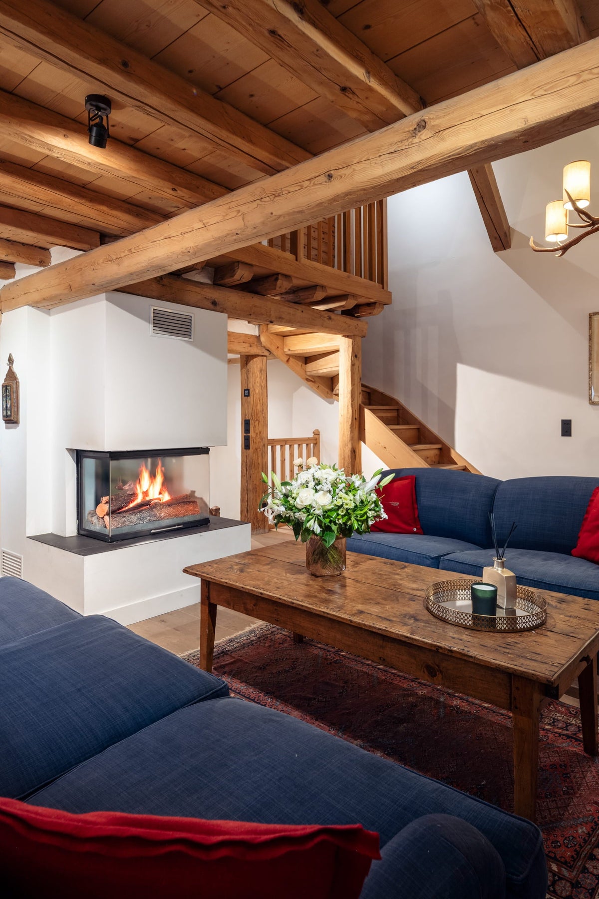 A cozy, rustic chalet-style living room featuring a large blue sectional sofa with red accent pillows, a matching blue sofa, a wooden coffee table, a fireplace, and exposed wooden ceiling beams.