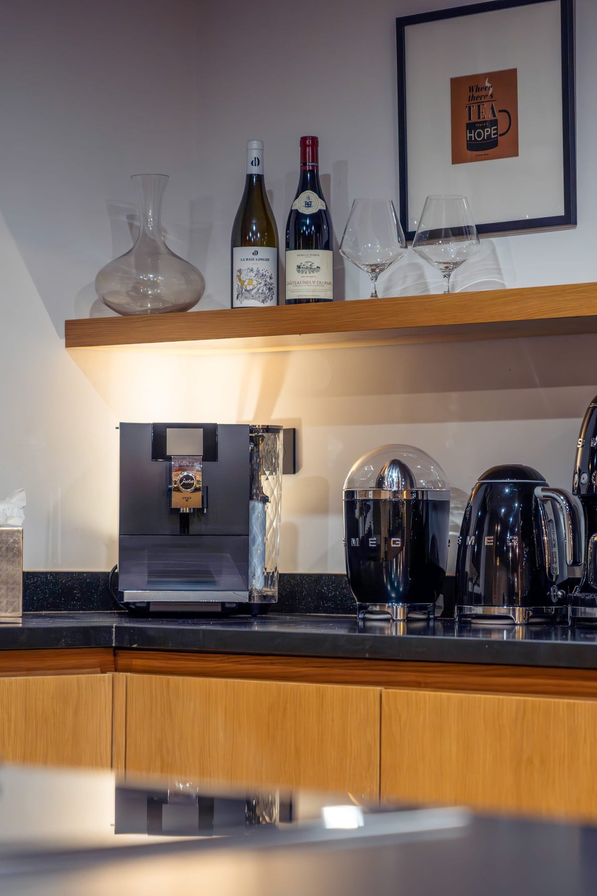 A modern kitchen countertop setup with black appliances including a coffee machine, kettle, and blender, set against light wood cabinetry and a white wall with framed art.