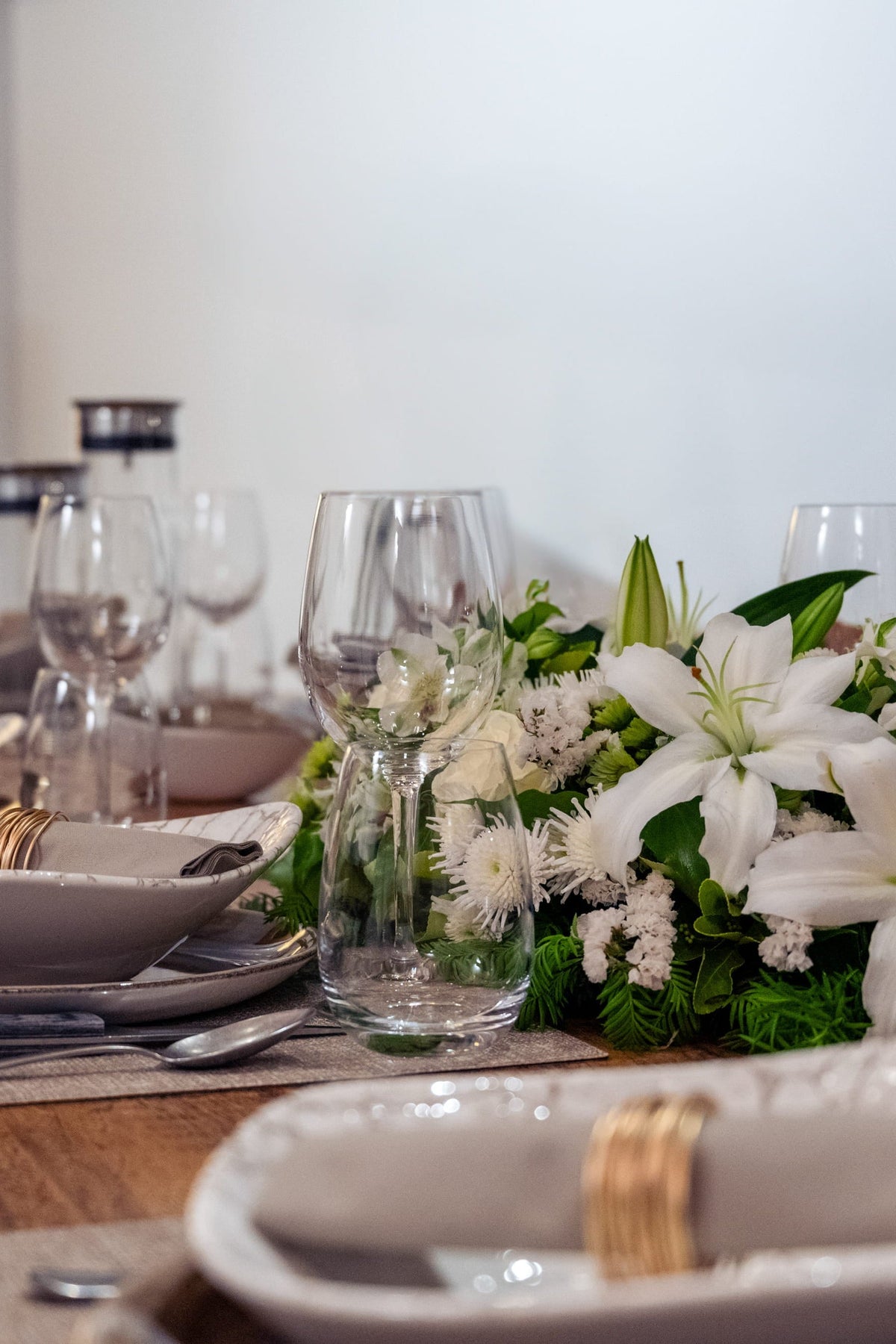 A beautifully set dining table with white lilies and green foliage centerpiece, white ceramic plates, gold ring napkins, and clear wine glasses in a warmly lit dining room.