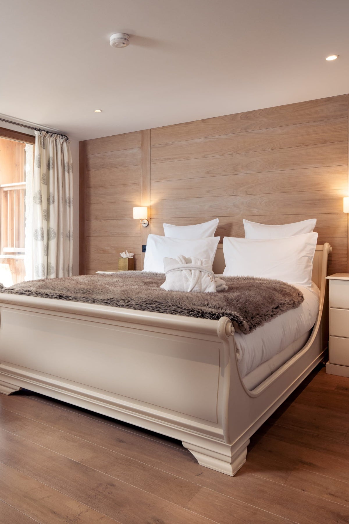 A beautifully styled bedroom featuring a cream-colored wooden sleigh bed with white linens and a brown faux-fur throw, set against a light wood-paneled wall. The room includes a matching cream nightstand, a wooden desk with a chair, a round mirror, and a large window with curtains.