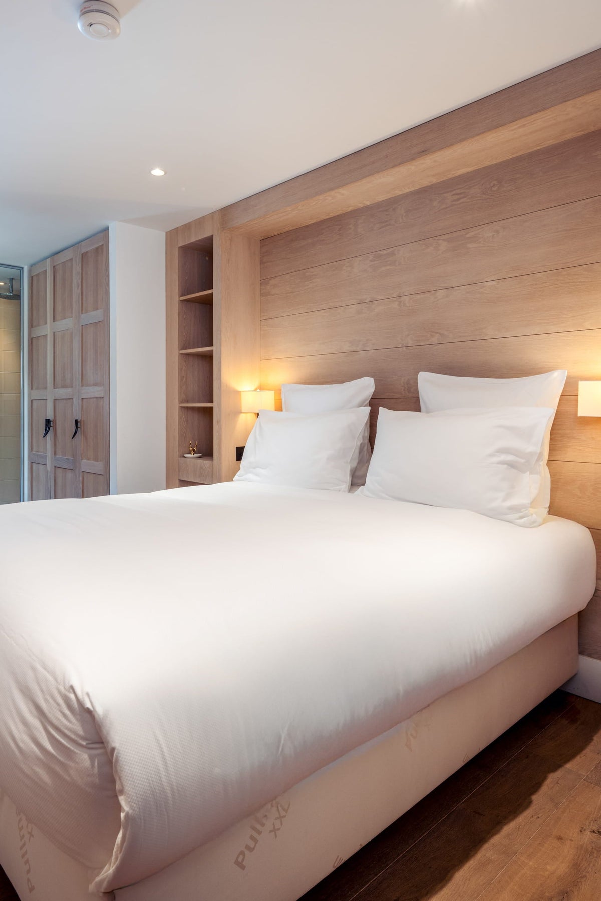 A modern bedroom featuring a large bed with white bedding, set against a light wood paneled headboard with built-in shelving and warm ambient lighting.