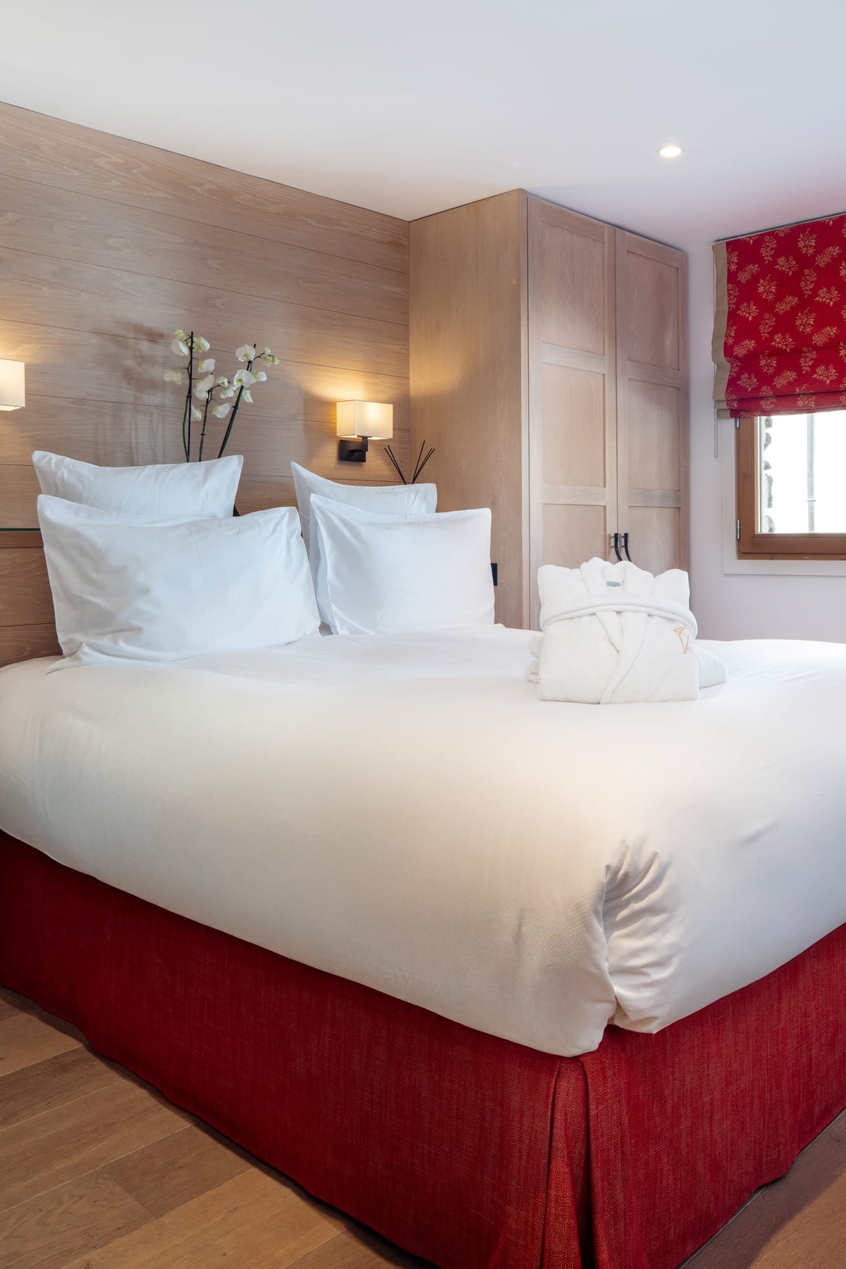 A double bed with white bedding and a red skirt in a modern bedroom with light wood furniture, a red patterned window valance, and a wooden chair.