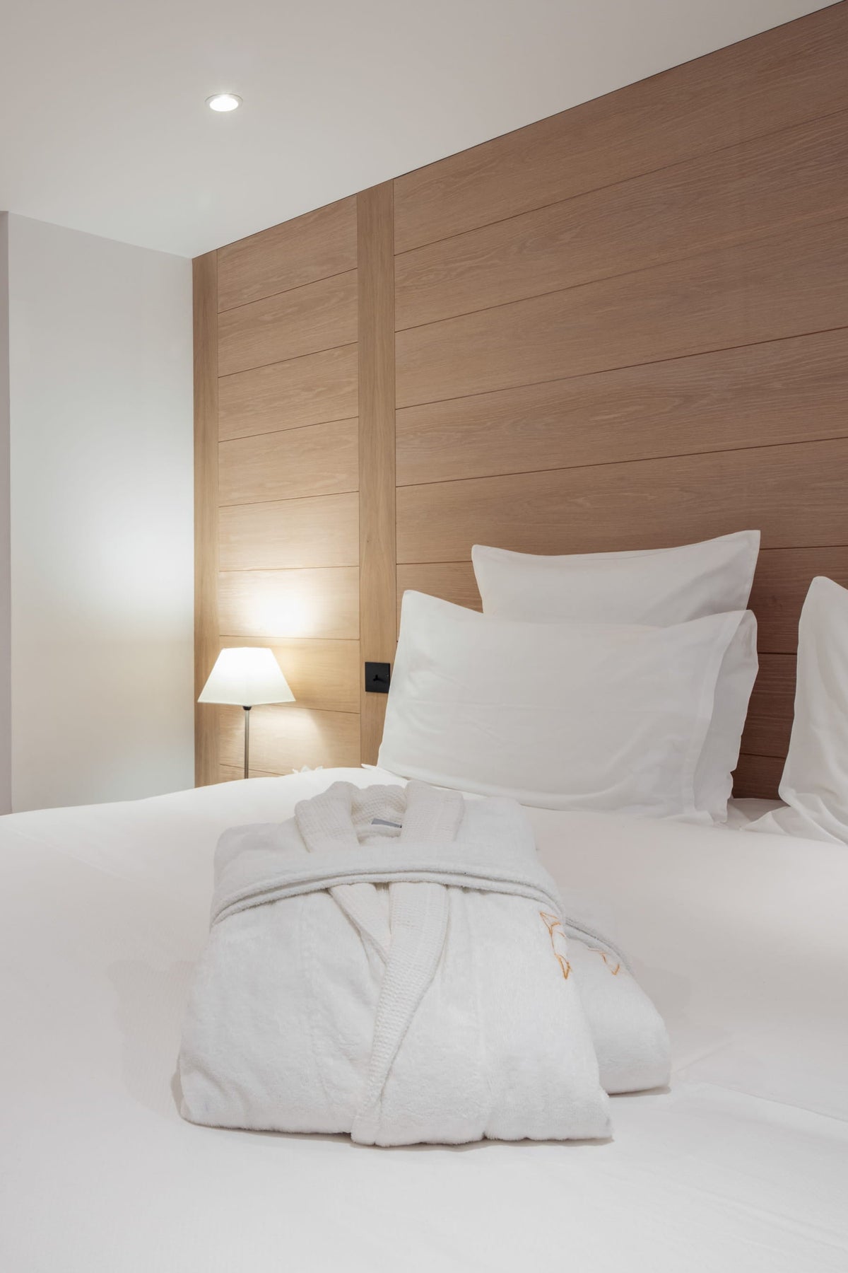 A modern bedroom featuring a neatly made bed with white linens and a folded white robe, set against a light wood headboard and matching wood wardrobes in a bright, contemporary room.
